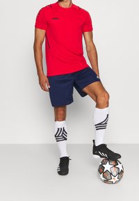 Hombre con camiseta deportiva roja, pantalones cortos azul marino, calcetas blancas con estampado y zapatillas negras, equilibrando un balón de fútbol negro y blanco con un pie.