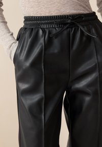 Black faux leather pants with a pleated design, featuring an elastic waistband and adjustable drawstring, side pockets, and smooth texture.