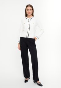 Cardigan de punto blanco con ribete negro y botones dorados, con dos bolsillos en el pecho, combinado con pantalones de pierna ancha negros y zapatos negros brillantes.