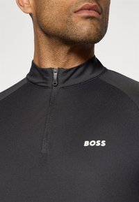 Svart atletisk zip-up topp laget av teksturert materiale, med høy krage og en liten hvit "BOSS"-logo på venstre side.