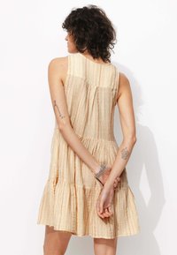 Robe sans manches à bretelles en beige avec des rayures verticales. Présente une jupe à volants et un détail froncé au dos pour plus de texture.