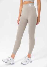 Beige bordázott leggings, magas derékkal, fehér logóval a derékpánton. Fehér sportcipővel viselve, szürke kiegészítőkkel.