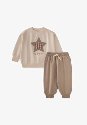 Kremasti duks s flasterom u obliku zvijezde u smeđem karo uzorku i tekstom "All of us star", upareno sa smeđim hlačama s elastičnim pojasom i manžetama.