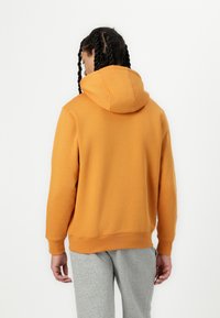 Sweatshirt laranja com capuz e um corte descontraído, apresentando um bolso canguru e pulsos em ribana. Usado com calças de moletom cinzas, mostrado pela parte de trás.