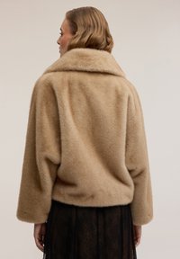 Giacca in pelo sintetico beige con ampio colletto, spalle scese e vestibilità morbida. Texture morbida, design leggermente corto, senza chiusure visibili.