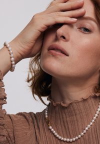 Naszyjnik z pereł z złotym zapięciem, noszony na teksturowanej brązowej bluzce z falbaną przy dekolcie. Na nadgarstku bransoletka z pasujących pereł.