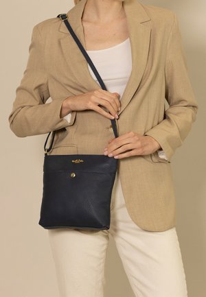 Navy leather crossbody bag with textured finish, gold hardware accents, and adjustable strap, held in front of a beige blazer.