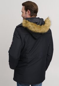 Black padded jacket with a fur-trimmed hood, featuring a water-resistant exterior. Includes logo patch on the sleeve and side pockets.