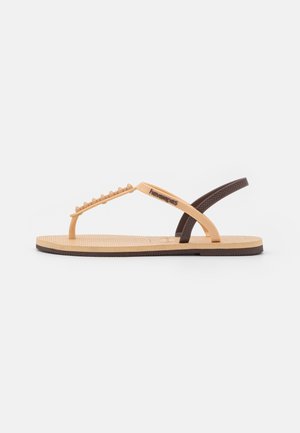Tan rubber flip-flops with a textured footbed, brown sole, and contrasting straps featuring decorative peaks and logo.