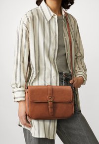 Brun læder crossbody taske med klaplukning, udstyret med en guldtonet trykknap og en aftagelig skulderrem, præsenteret mod en stribet skjorte.