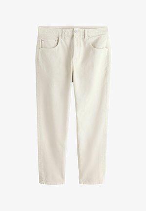 Detail ľavej ruky osoby odpočívajúcej vedľa svetlo béžových denimových džínsov s predným vreckom a nitovými detailmi, oblečená v bielom rebrovanom tričku.