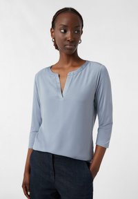 Lichtblauwe blouse met lange mouwen en een V-hals. Gemaakt van een soepele stof. Heeft een relaxte pasvorm en een strak, minimalistisch ontwerp.