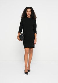 Abito a coste nero con maniche a tre quarti, scollatura alta e silhouette aderente. Abbinato a una piccola borsa nera e scarpe ballerine.