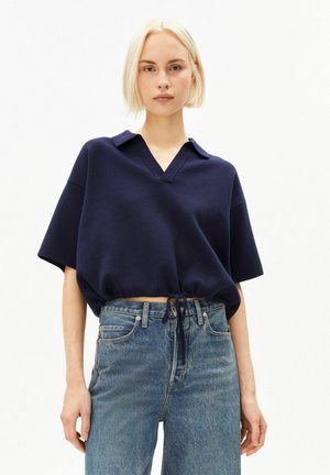 Junge Frau mit kurzen blonden Haaren, die ein marineblaues kurzärmeliges Oberteil mit Kragen und hoch taillierte blaue Jeans vor einem schlichten weißen Hintergrund trägt.