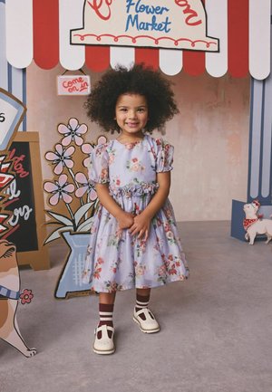 Šypsanti mergina su garbanotais plaukais, dėvinti gėlėtą suknelę ir baltus batus, stovi prie gėlių turgavietės prekystalo su animacinių šunų išpjovomis.