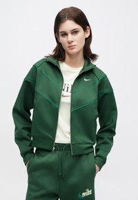 Νέος με κοντά καστανά μαλλιά φοράει πράσινη φόρμα Nike και λευκό T-shirt Nike, στέκεται με τα χέρια στις τ σέπες μπροστά σε απλό φόντο.