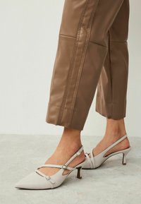 Světlé šedé slingback podpatky s špičatou špičkou, nastavovacími řemínky a malým blokovým podpatkem. V páru s hnědými zkrácenými kalhotami.