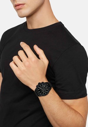 Montre chronographe noire avec un bracelet texturé et plusieurs cadrans, portée sur un poignet avec une chemise à manches courtes noire.