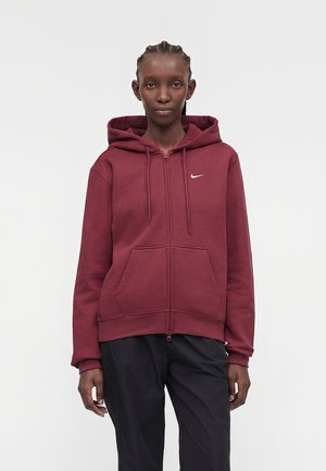 Kobieta z warkoczami, ubrana w bordową bluzę z kapturem na zamek błyskawiczny z białym logo Nike i czarne spodnie, stoi na jednolitym jasnym tle.