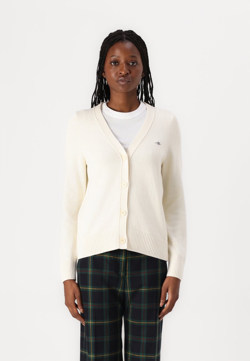 Cardigan beige con scollo a V e sei bottoni, con polsini e orlo a coste. Indossato sopra una maglietta bianca e abbinato a pantaloni a quadri.