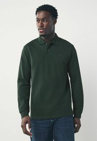 Dunkelgrünes Langarm-Polohemd mit Kragen, zwei Knöpfen und einem kleinen gestickten Logo. Kombiniert mit blauen Jeans.