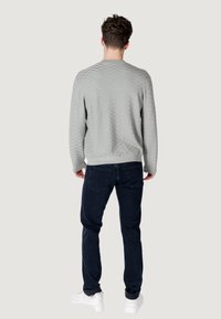 Grijze, gestructureerde trui met ruitpatroon; aansluitende manchetten en tailleband. Gedragen met donkere spijkerbroeken en witte sneakers, gezien vanuit de achterkant.