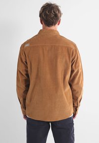 Chemise en velours côtelé marron avec des rayures verticales, poignets à boutons et ourlet arrondi. Présente une étiquette sur l'empiècement. Vue arrière présentée.