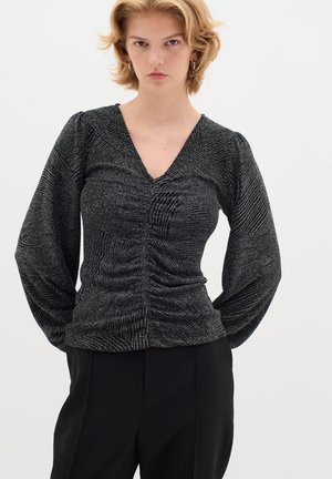 Svart texturerad långärmad topp med V-ringning, med rynkade detaljer över framsidan och voluminösa ärmar. Bärs med svarta byxor.