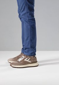 Sneakers in pelle scamosciata marrone con dettagli bianchi, design con lacci e suola in gomma testurizzata, abbinati a pantaloni slim fit blu.