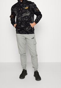 Czarna bluza z wzorem, z kieszenią na froncie i żółtym logo, zestawiona z szarymi spodniami dresowymi cargo z boczną kieszenią i ściągaczami na kostkach.