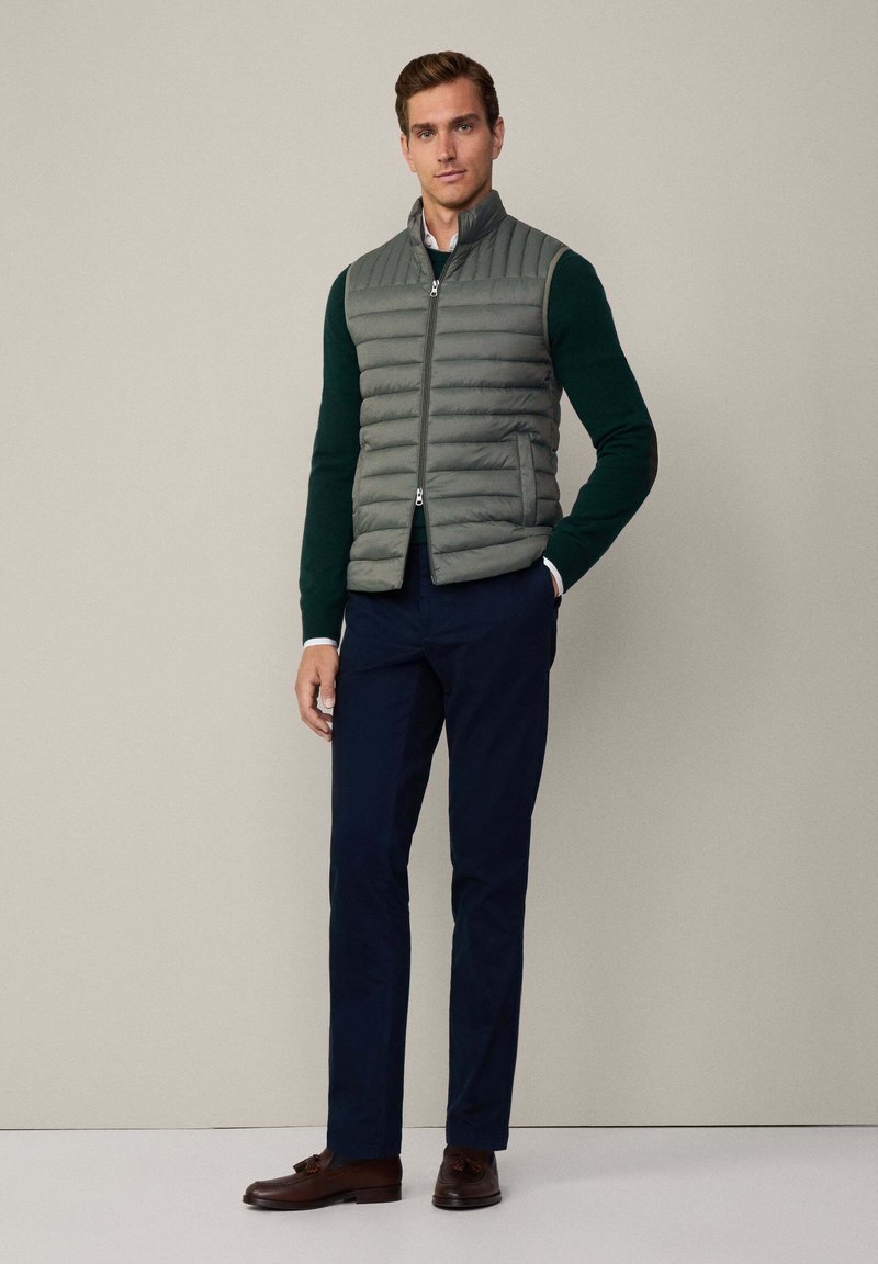Grau gepolsterte Weste mit vertikalen Nähten, mit Reißverschluss, getragen über einem dunkelgrünen Pullover, kombiniert mit dunklen Hosen und braunen Schuhen. Einfaches Design.