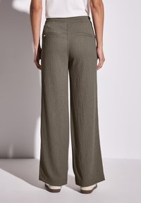 Wijde, olijfgroene broek met een gestructureerde stof; beschikt over twee achterzakken en een gladde tailleband, gecombineerd met lichte schoenen.