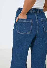 Dunkelblaue Jeans mit geradem Bein, elastischem Bund und zwei aufgesetzten Taschen auf der Rückseite. Strukturiertem Stoff mit sichtbaren Nähten.
