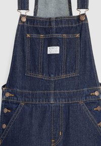 Pantalones de mezclilla azul oscuro con tirantes ajustables, bolsillo frontal en el pecho con etiqueta de Levi Strauss, botones laterales y detalles de costura amarilla.