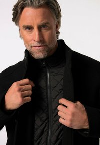 Abrigo exterior negro con un cuello alto, que cuenta con un forro interior acolchado. Textura suave y cierre con cremallera en la parte delantera. El modelo ajusta el abrigo.