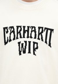 Gros plan d'un pull crème avec le texte noir tricoté « CARHARTT WIP » sur la poitrine en police grasse et légèrement incurvée.