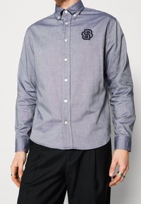 Ljusblå skjorta med knappar, i bomullsblandning, med rundad nederkant och marinblå broderad logotyp på bröstet. Har klassisk krage och knäppning.