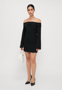 Svart av-shoulder kjole med lange ermer, tettsittende silhuett og glatt tekstur. Kombinert med en hvit stoffveske og svarte spisse hæler.