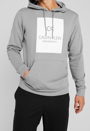 Mężczyzna w jasnoszarej bluzie z kapturem Calvin Klein Performance z przednią kieszenią typu kangur i czarnych spodniach, ręka w kieszeni.