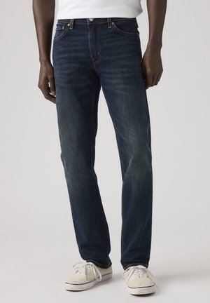 Uomo con jeans blu scuro a gamba dritta e sneakers bianche su uno sfondo uniforme.
