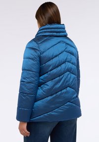 Chaqueta acolchada en material azul oscuro brillante, con un cuello alto y un patrón de acolchado en forma de chevrón. Textura suave con costuras visibles.