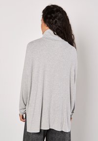 Helles graues, locker sitzendes Oberteil mit hohem Kragen und langen Ärmeln. Weiche Textur, gespaltene Saumlinie und entspannte Silhouette. Der Rücken ist schlicht und ohne sichtbare Details.