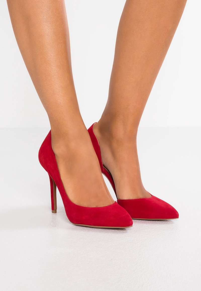 Femme portant des escarpins aiguilles en daim rouge, se tenant sur un sol blanc.