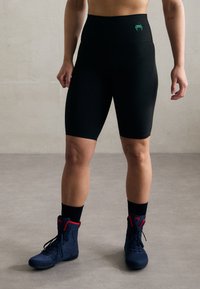 Pantaloni da ciclismo neri a vita alta realizzati in tessuto elasticizzato, con un logo verde. Abbinati a stivali alla caviglia blu con stringhe e accenti rossi, e calze nere.