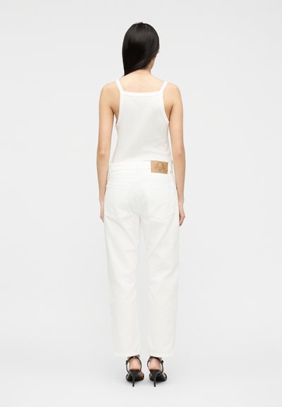 Γυναίκα με μακριά σκούρα μαλλιά, που φοράει λευκό αμάνικο τοπ, λευκό τζιν με δερμάτινο ταμπελάκι και μαύρα σανδάλια με ψηλό τακούνι, γυρισμένη με την πλάτη.