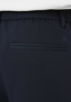 Ceinture de pantalon bleu marine foncé avec dos élastique et poche passepoilée unique, montrant la texture du tissu et une partie du logo sur le côté droit.