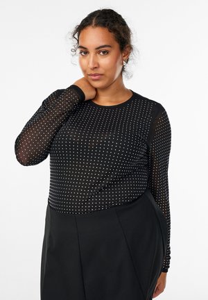 Black long-sleeve top with a sheer mesh fabric dotted with white patterns, rounded neckline, and fitted design, paired with high-waisted black pants.
