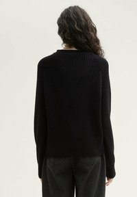 Zwart gebreid sweater met een gestructureerde weving, ronde hals, en geribbelde boorden en zoom, gecombineerd met losse donkere jeans. Zicht van achter.