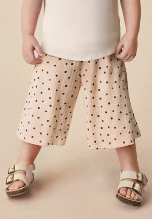 Barn iført beige shorts med sorte prikker, hvid ærmeløs top og hvide sandaler med guldfarvede spænder, stående på en neutral baggrund.