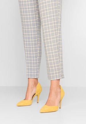 Gule ruskind høje hælede pumps med spids tådesign. Parret med grå ternede korte bukser med et blå og gult ternmønster.